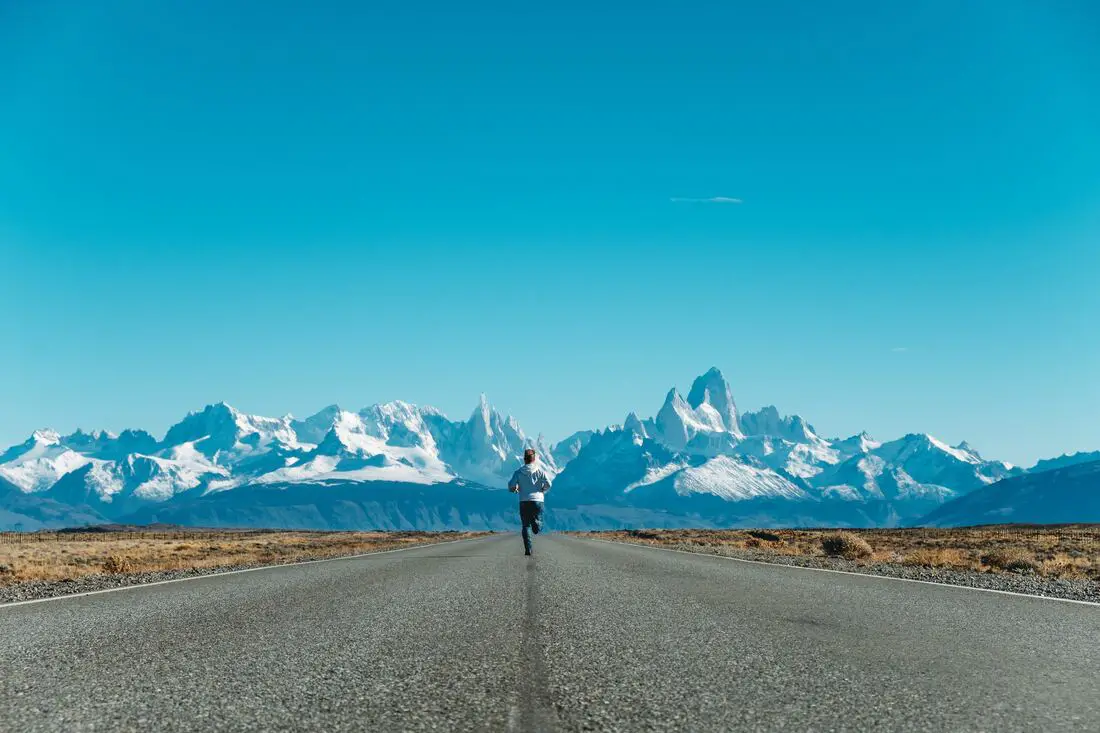 man running in the middle of road towards snow capped mountains